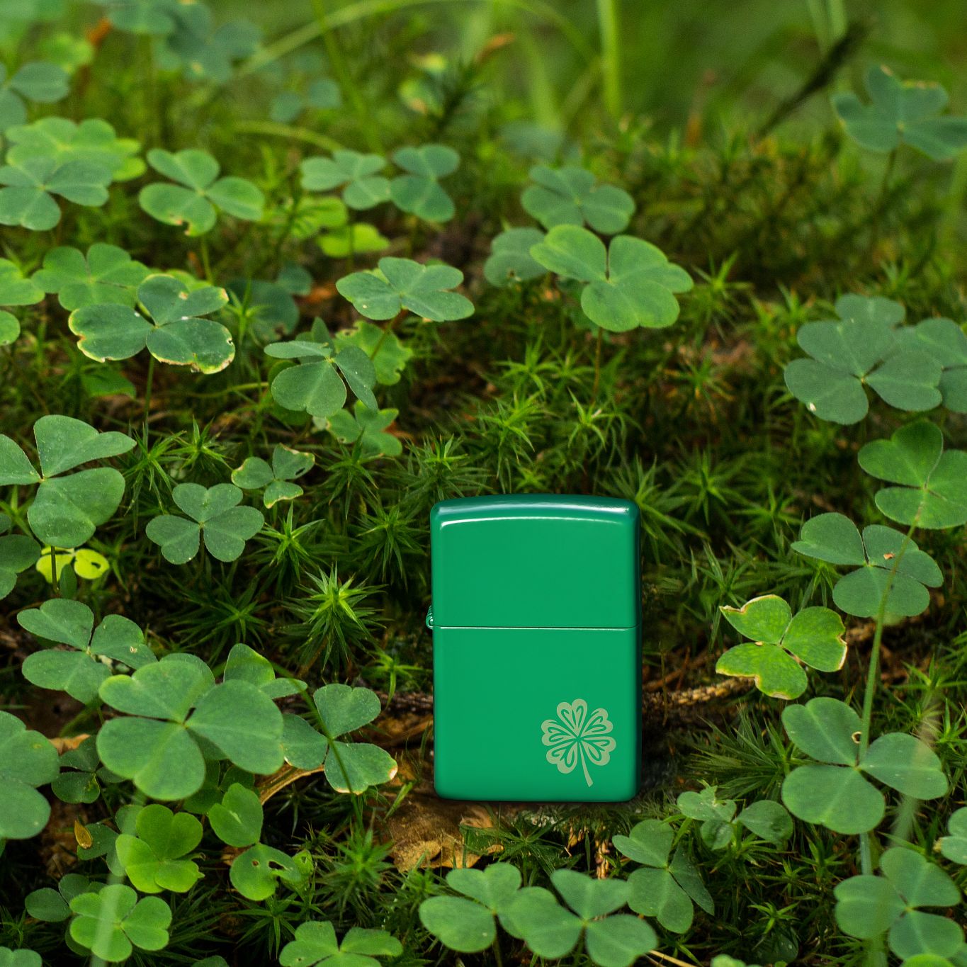 Lifestyle image of Zippo Lucky Clover Design Grass Green Matte Windproof Lighter standing in a bed of clover with some twigs and leaves.