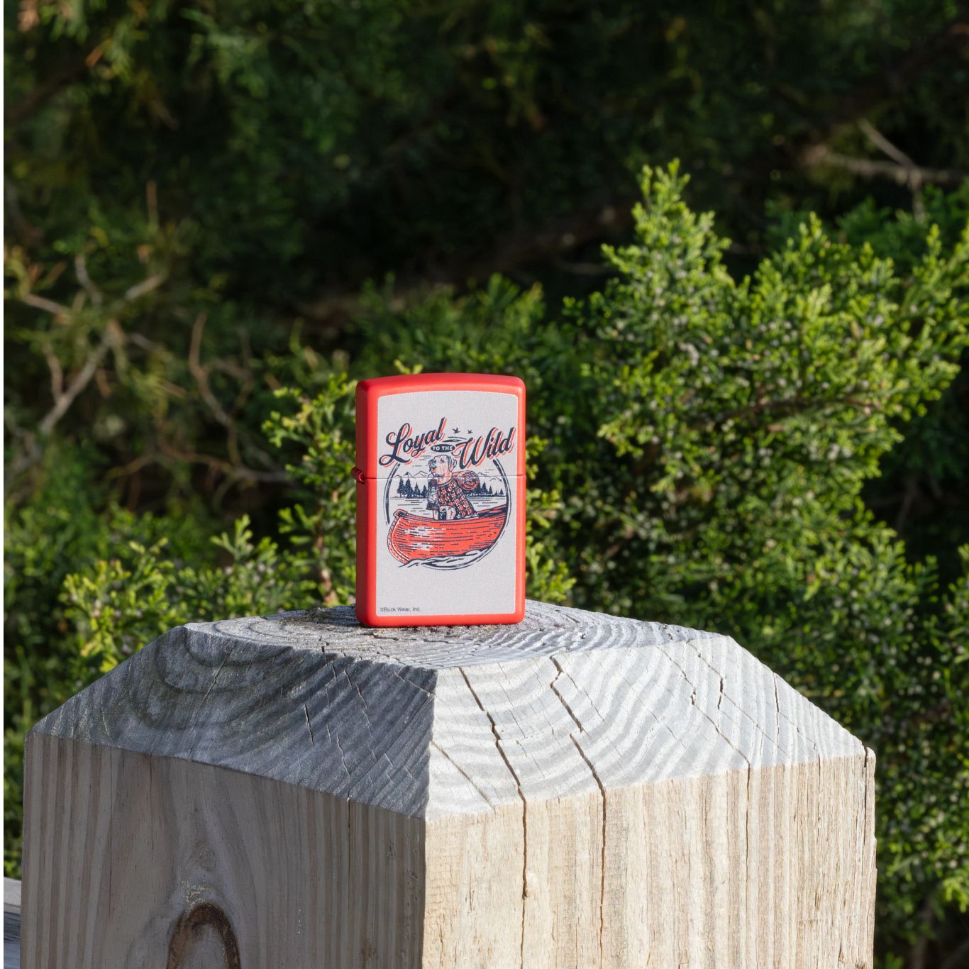 Lifestyle image of Zippo Buck Wear™ Loyal to the Wild Design Red Matte Windproof Lighter standing on a porch post with a wooded scene in the background.