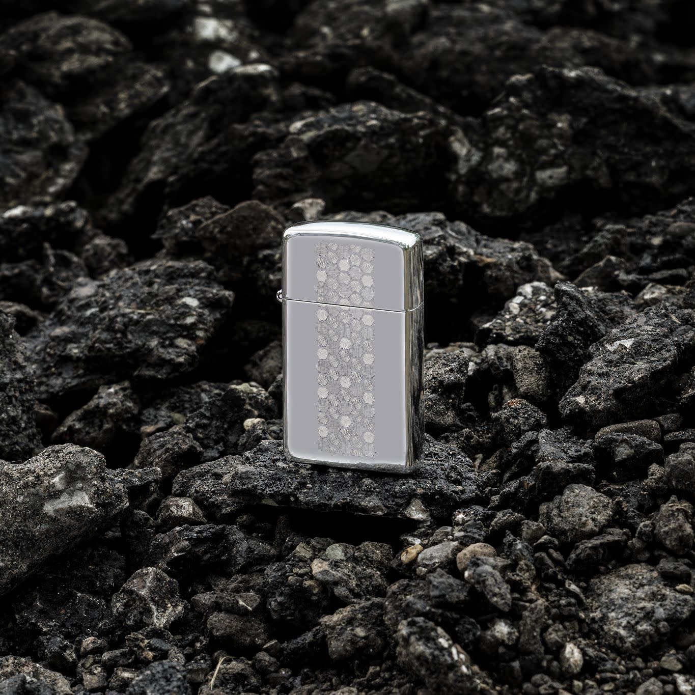 Lifestyle image of Zippo Slim® Honeycomb Design High Polish Chrome Windproof Lighter standing on dark, loose rocks.