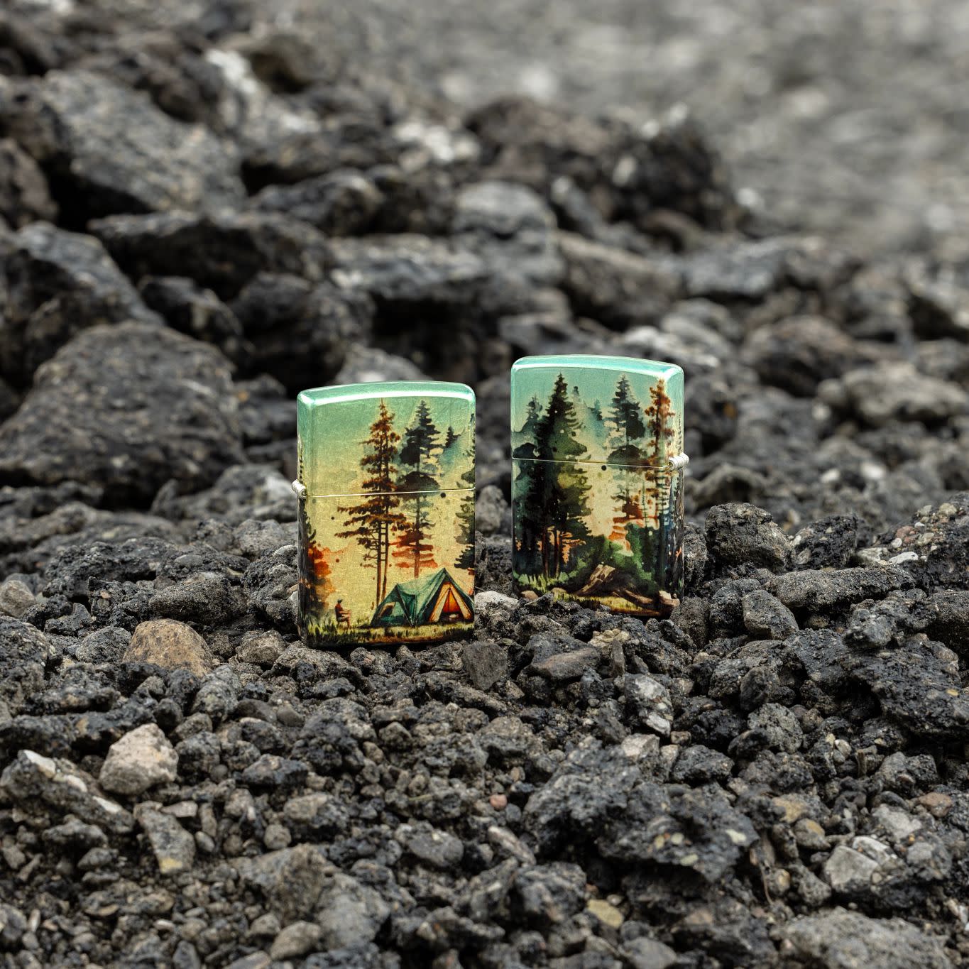 Lifestyle image of two Zippo Off the Grid Design 540 Tumbled Brass Windproof Lighters standing on dark, loose rocks; one showing the front of the lighter and the other showing the back.