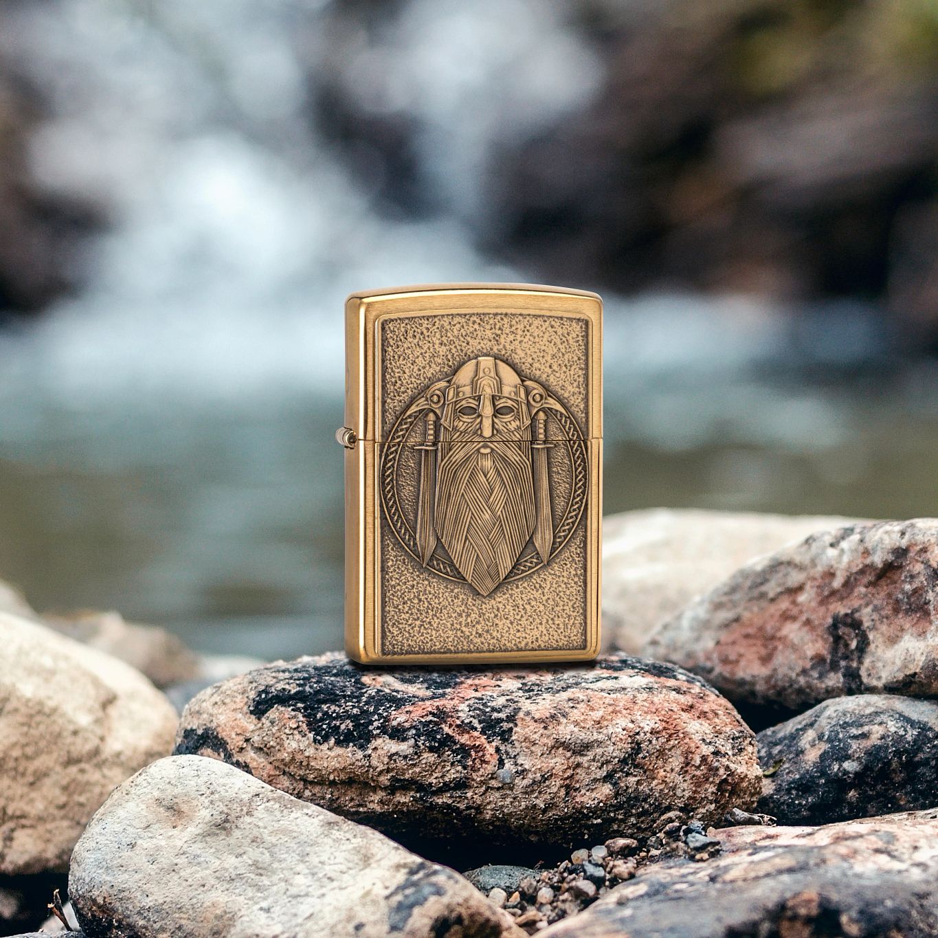 Lifestyle image of Zippo Odin Emblem Brushed Brass Windproof Lighter standing on river rocks with a blurred waterfall and large rocks in the background.