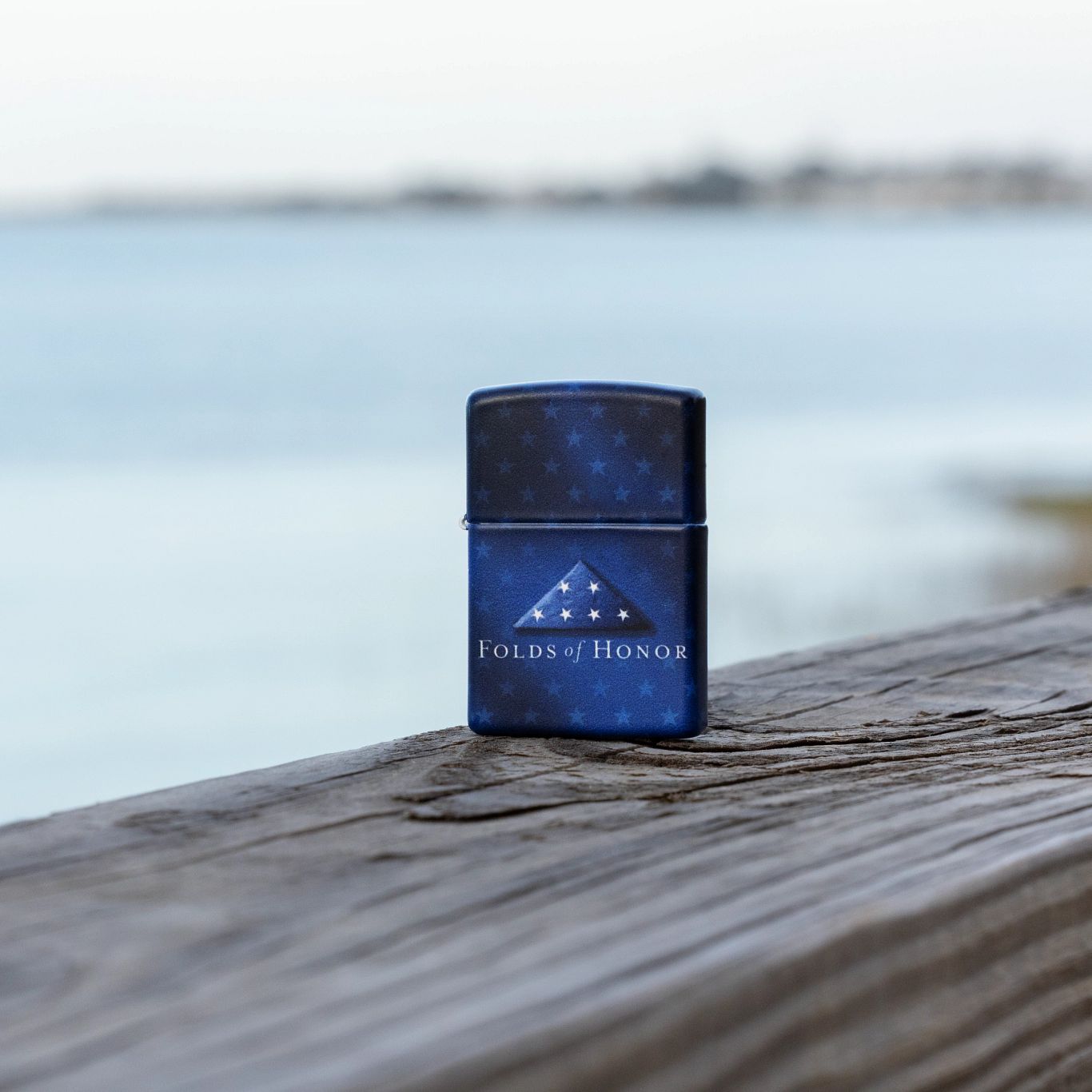 Lifestyle image of Zippo Folds of Honor Star-Studded Design 540 Matte Windproof Lighter standing on a wooden rail with a seascape scene in the background.