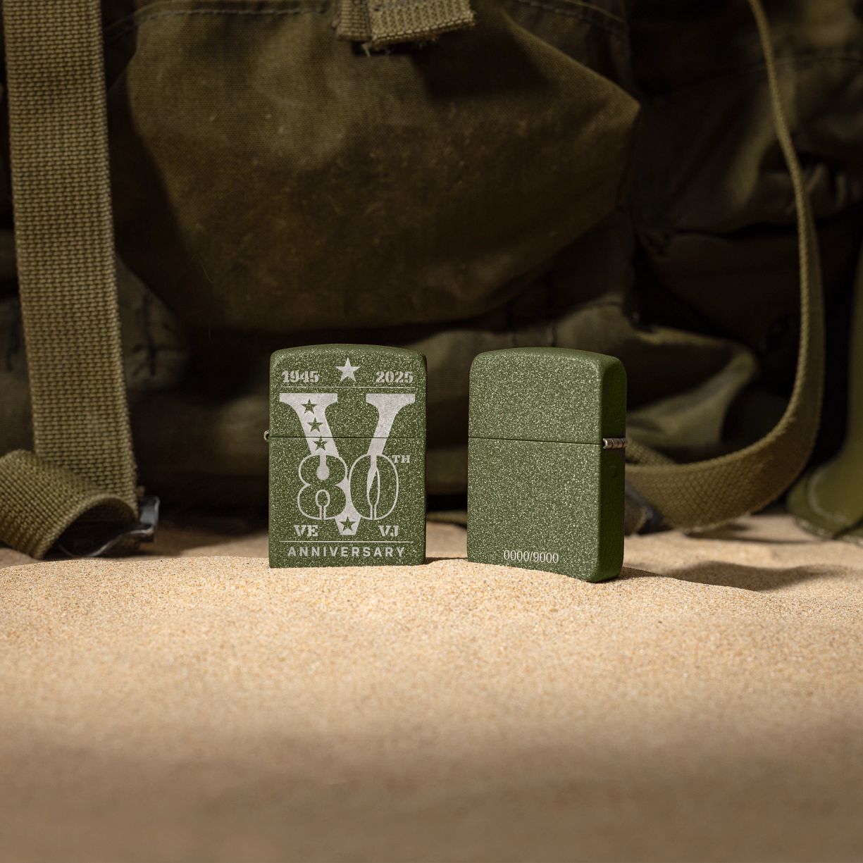 Lifestyle image of two Zippo VE/VJ 80th Anniversary Collectible Steel Windproof Lighters, one showing the front of the lighter and the other showing the back, standing in front of a military bag.