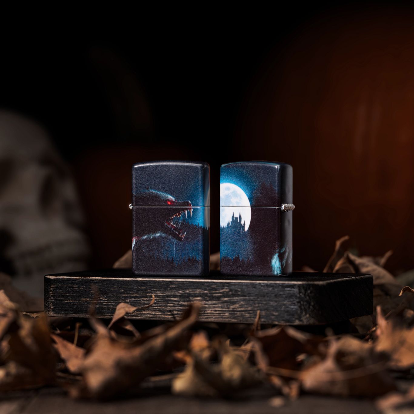 Lifestyle image of two Zippo Horror Wolf 540 Matte Windproof Lighters; one showing the front of the lighter while the other shows the back and hinge side of the lighter. They are both standing on a dark wooden block surrounded by brown leaves with a skull in the background.