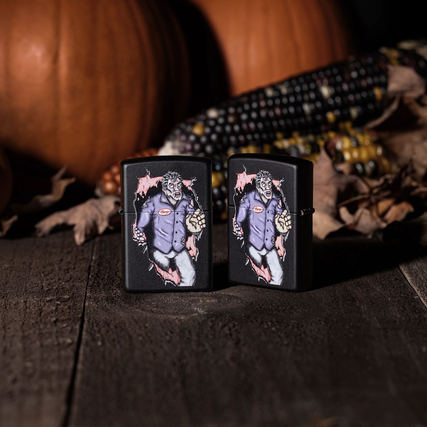 Lifestyle image of two Zippo Zombie Escape Design Black Matte Windproof Lighters; one showing the front of the lighter while the other shows the back and hinge side of the lighter. They are both standing on a wooden floor with pumpkins, leaves, and corn in the background.