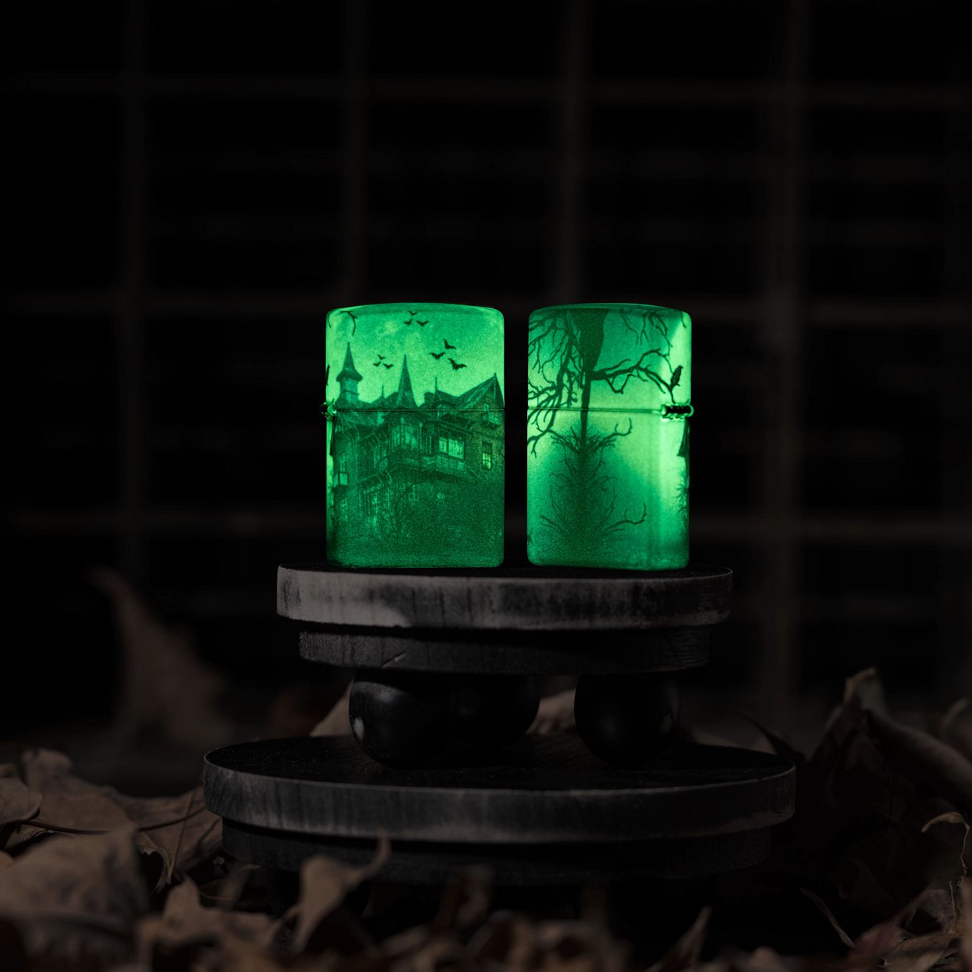 Lifestyle image of two Zippo Horror House Glow in the Dark Matte Windproof Lighters glowing in the dark; one showing the front of the lighter while the other is showing the back and hinge side of the lighter. They are both standing on a tiered wooden riser surrounded by brown leaves with a dark background.