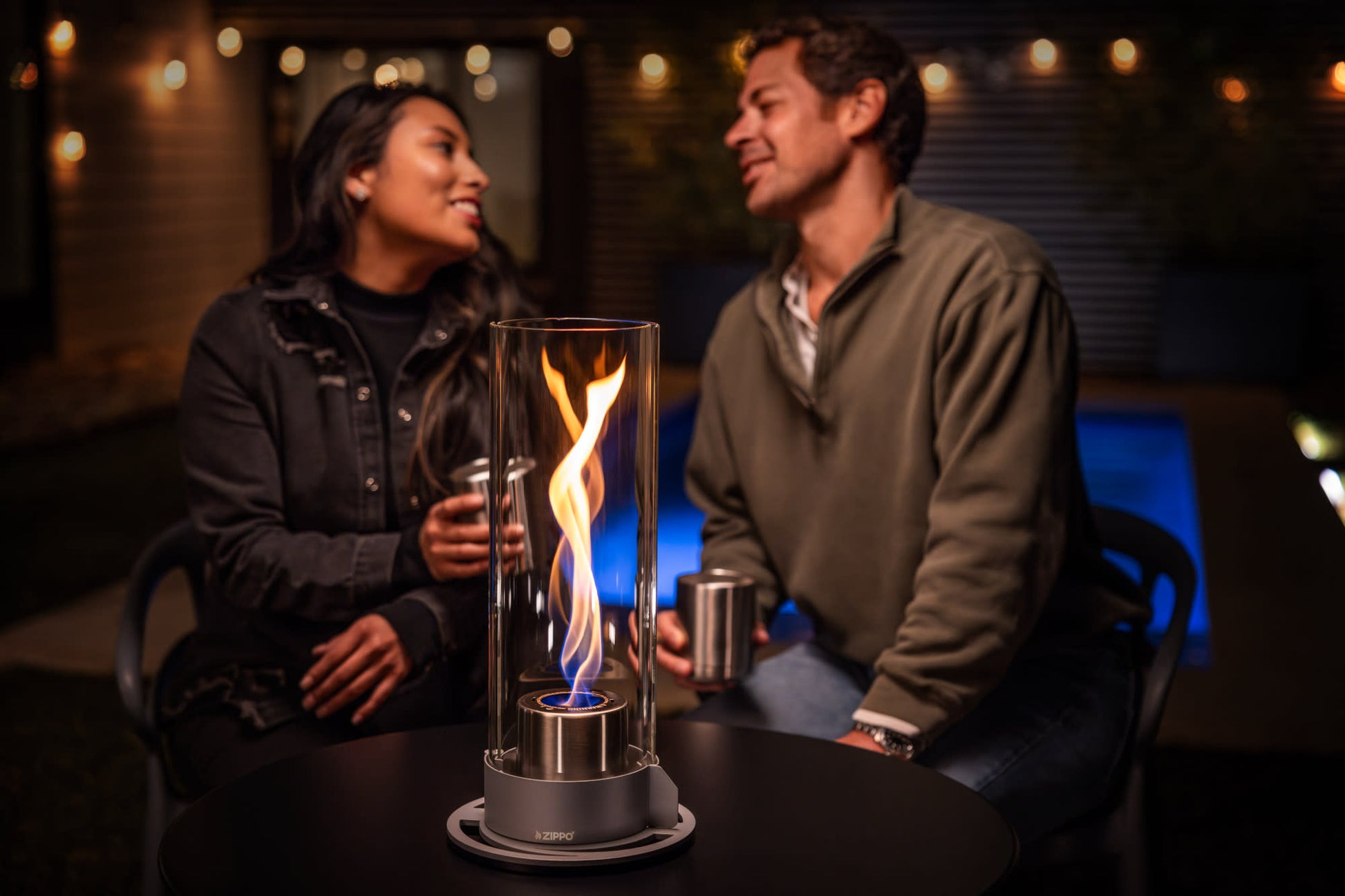 Lifestyle image of the Zippo FlameScapesâ„¢ Spiral Fire Feature XL in black, lit on a table with a cityscape and couple in the background.