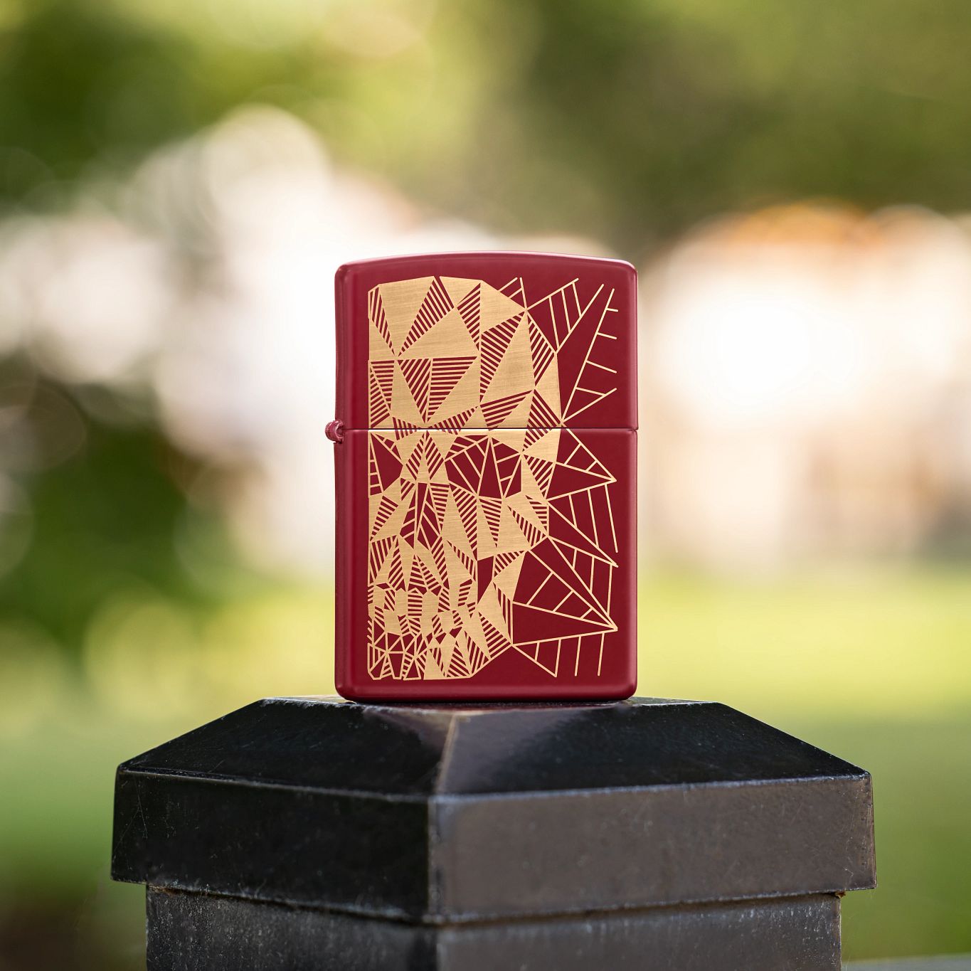 Lifestyle image of Zippo Geometric Skull Design Merlot Windproof Lighter standing on top of a black metal railing post showing the front of the lighter with an out of focus city park as the background.