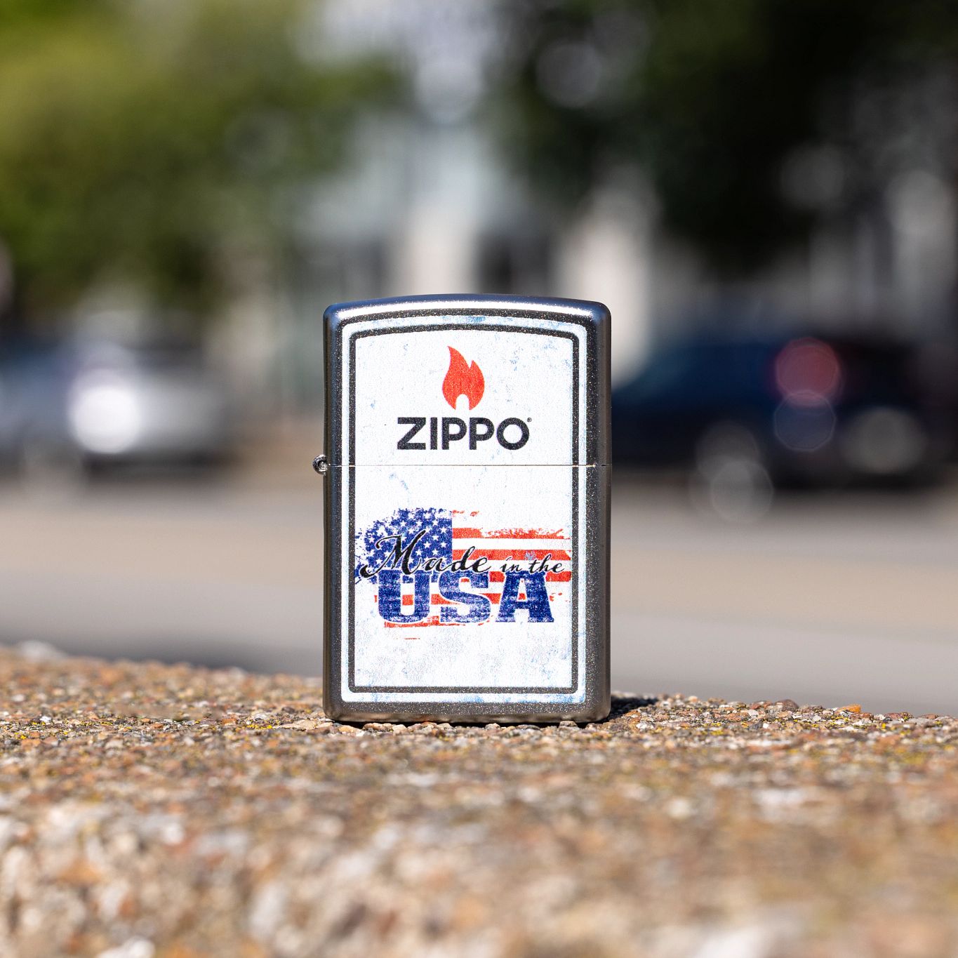 Lifestyle image of Zippo Made in the USA Design Satin Chrome Windproof Lighter standing on a curb showing the front of the lighter with a blurred building, street, cars, and trees as the background.