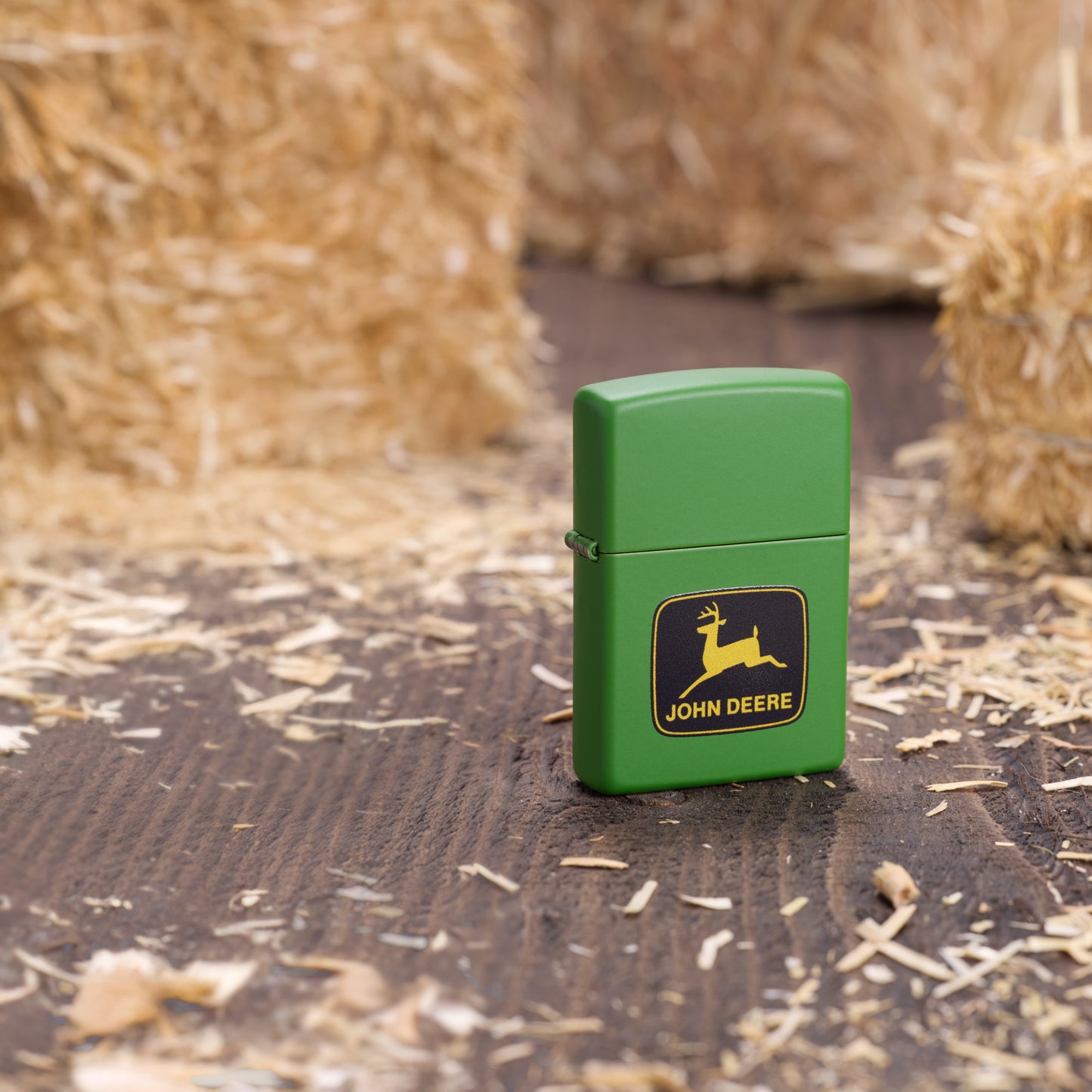 Lifestyle image of Zippo John Deere Logo Design Moss Green Windproof Lighter standing on a wooden floor surrounded by hay bales.