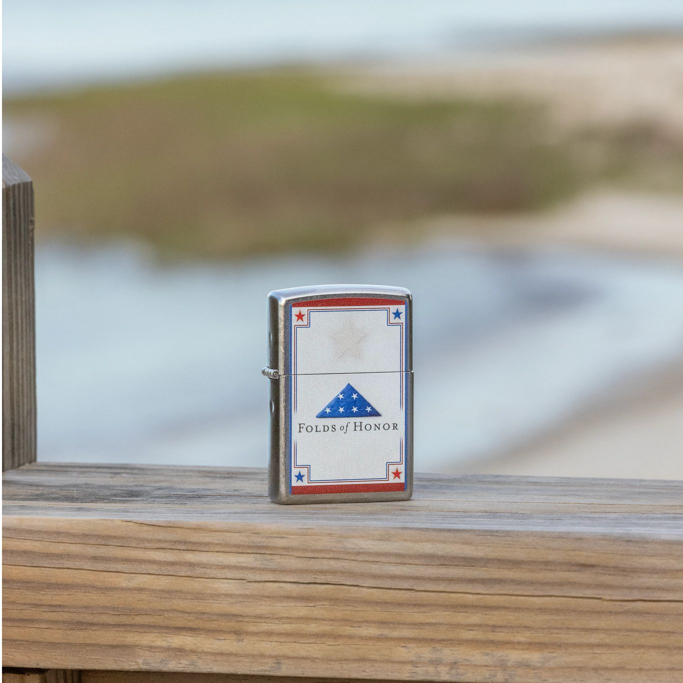 Lifestyle image of Zippo Folds of Honor Logo Design Street Chrome Windproof Lighter standing on a wooden rail with a blurred seascape scene in the background.