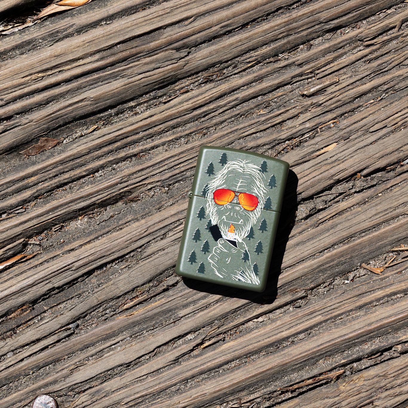 Lifestyle image of Zippo Zipsquatch Design Green Matte Windproof Lighter laying on a worn wooden walkway.