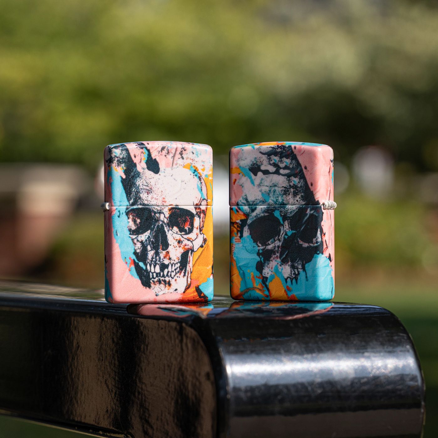 Lifestyle image of two Zippo Graffiti Skull Design 540 Matte Windproof Lighters; one showing the front of the lighter and the other showing the back and hinge side of the lighter, both standing on the armrest of a black metal park bench with an out of focus city park background.