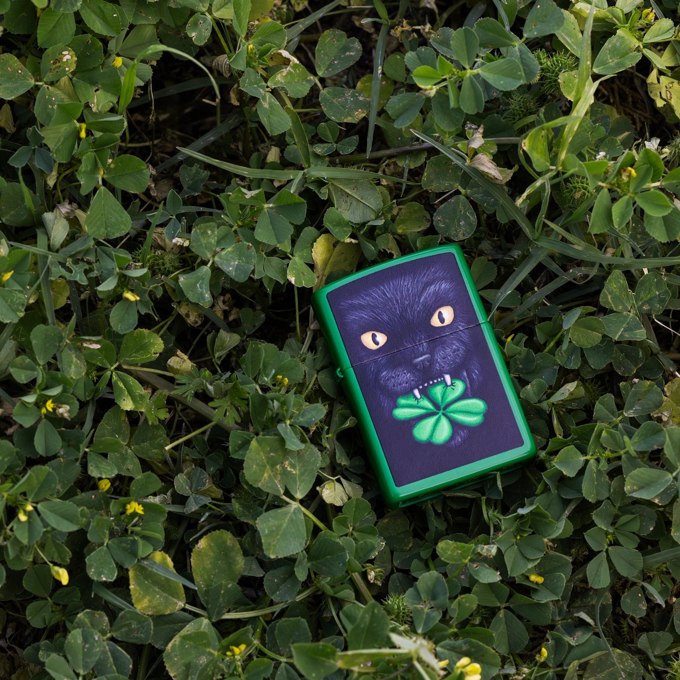 Lifestyle image of Zippo Feline Lucky Design Grass Green Matte Windproof Lighter laying in a pile of clovers.