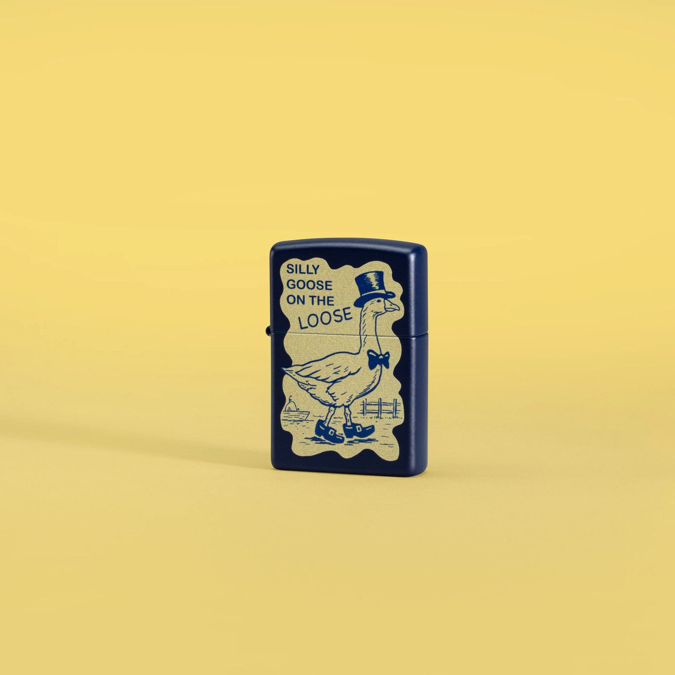Lifestyle image of Zippo Silly Goose Design Navy Blue Matte Windproof Lighter set against a yellow background.