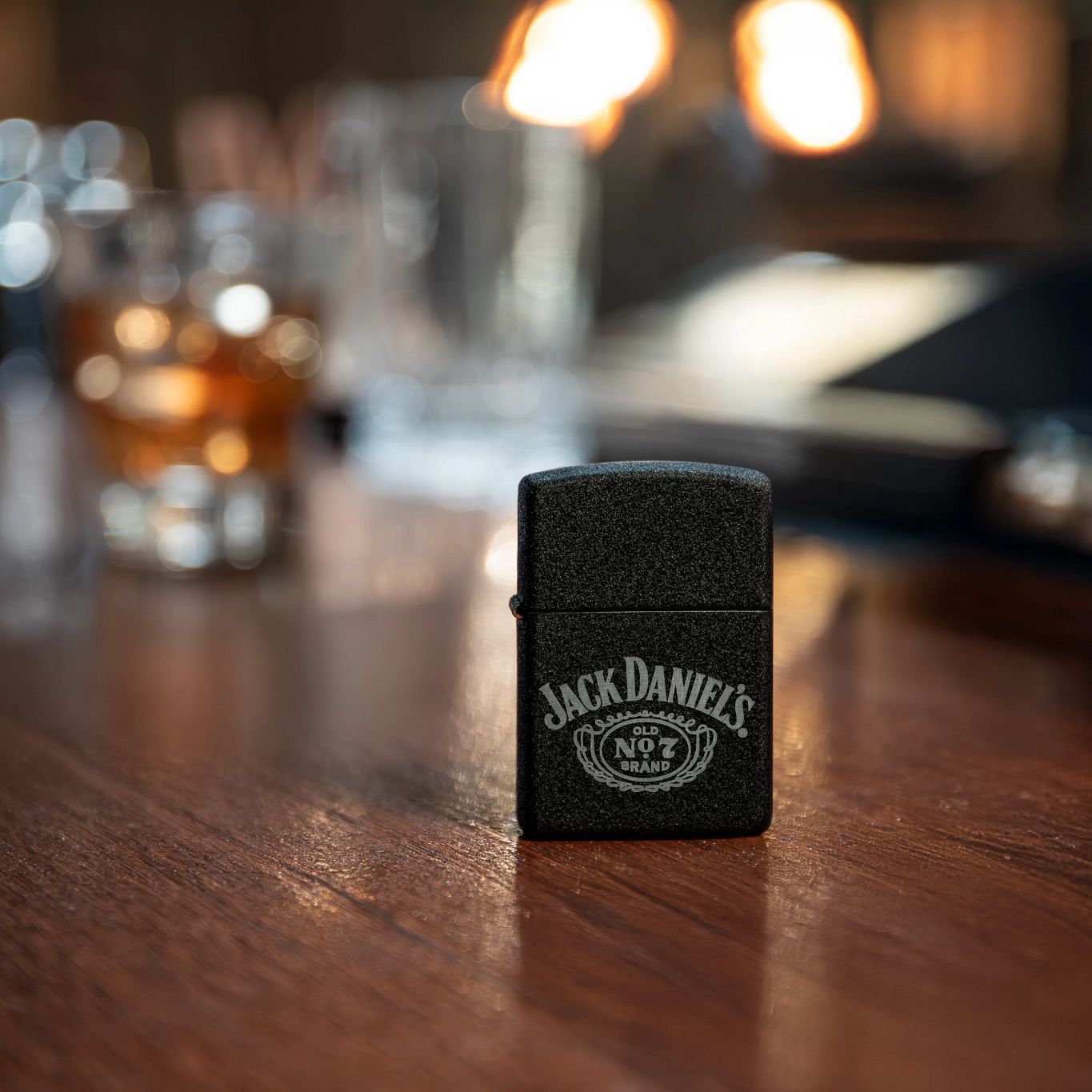 Lifestyle image of Zippo Jack Daniel's® No. 7 Design 1941 Replica Black Crackle® Windproof Lighter standing on a bar next to a glass of whiskey with the pub blurred in the background.