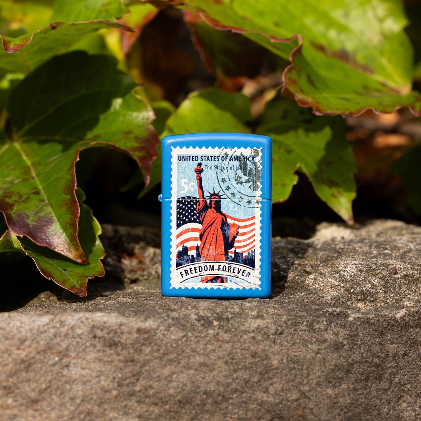 Lifestyle image of Zippo Stamp of Liberty Design Sky Blue Matte Windproof Lighter standing on a rock showing the front of the lighter with green and dark brown leaves behind the lighter.