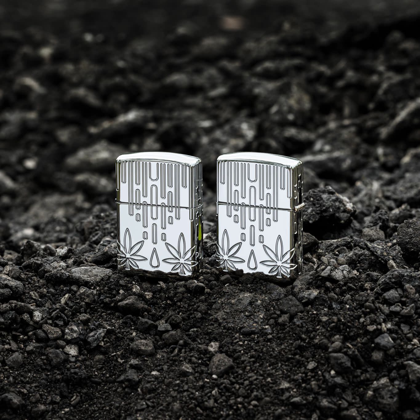 Lifestyle image of two Zippo Waxy Leaf Design Armor® High Polish Chrome Windproof Lighters standing on dark, loose rocks; one showing the front of the lighter and the other showing the back.
