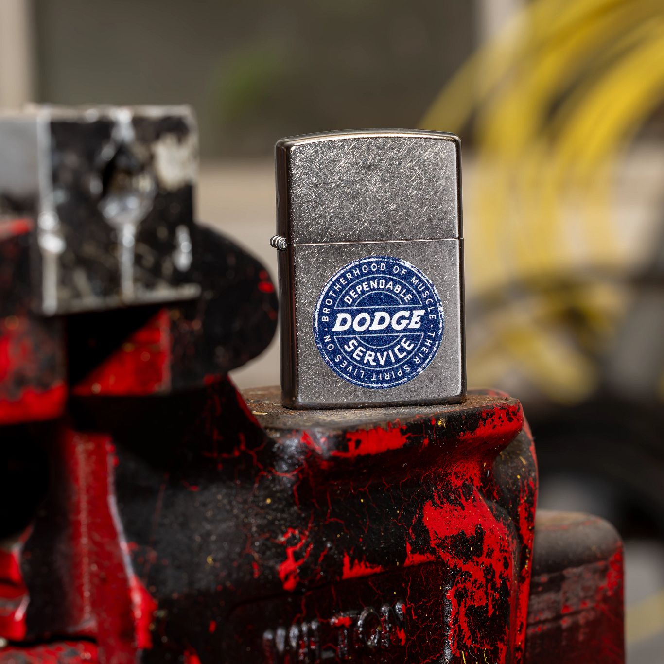 Lifestyle image of Zippo Dodge Service Sign Satin Chrome Windproof Lighter standing on an old mechanics pro vise with a blurred garage in the background.