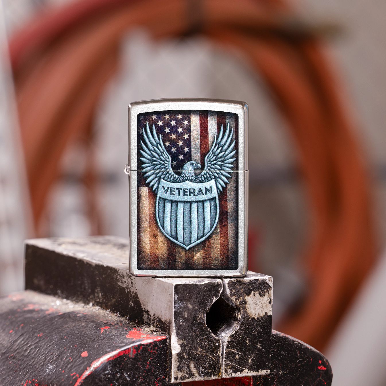 Lifestyle image of Zippo Veteran Crest Design Street Chrome Windproof Lighter showing the front of the lighter while standing on a well-used vise grip with a blurred garage as the background.