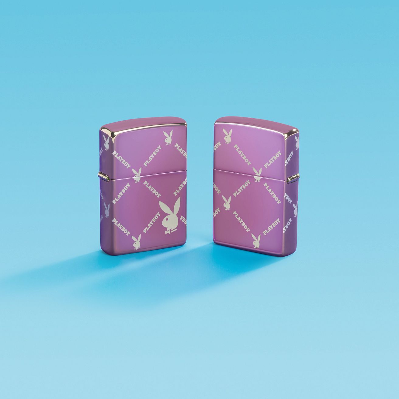 Lifestyle image of two Zippo Playboy Argyle Design High Polish Purple Windproof Lighters; one showing the front of the lighter and the other showing the back and hinge side of the lighter, both standing with a light blue background.