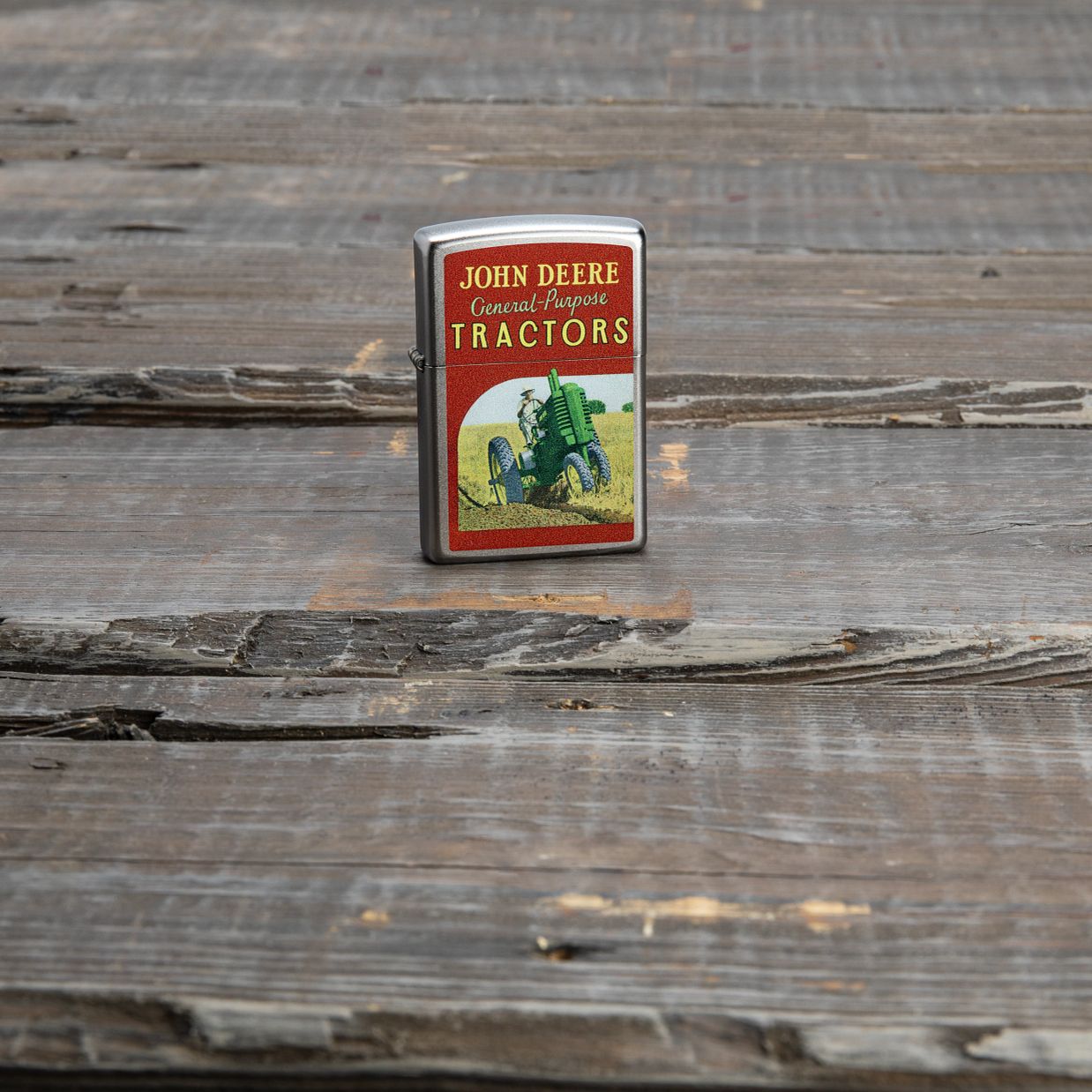 Lifestyle image of Zippo John Deere Vintage Ad Design Satin Chrome Windproof Lighter standing on a worn wooden porch floor.
