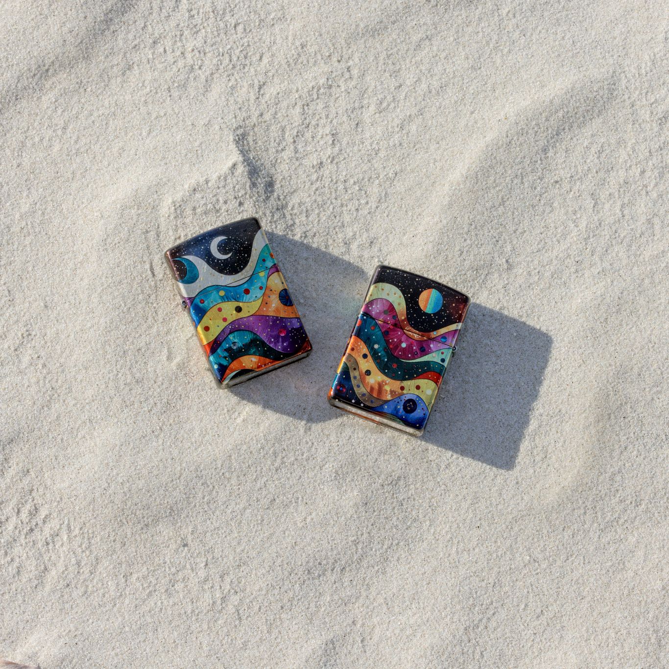 Lifestyle image of two Zippo Space Fusion Design 540 Tumbled Chrome Windproof Lighters, one showing the front of the lighter and the other showing the back, both laying in the sand.