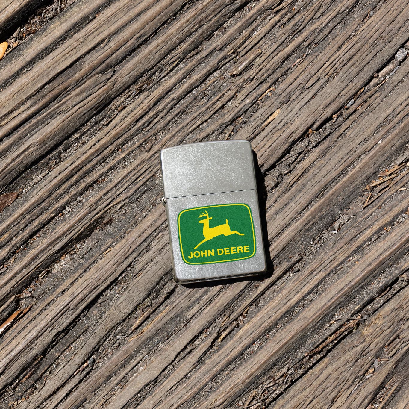 Lifestyle image of Zippo John Deere Leaping Deer Design Street Chrome Windproof Lighter laying on a worn wooden walkway.