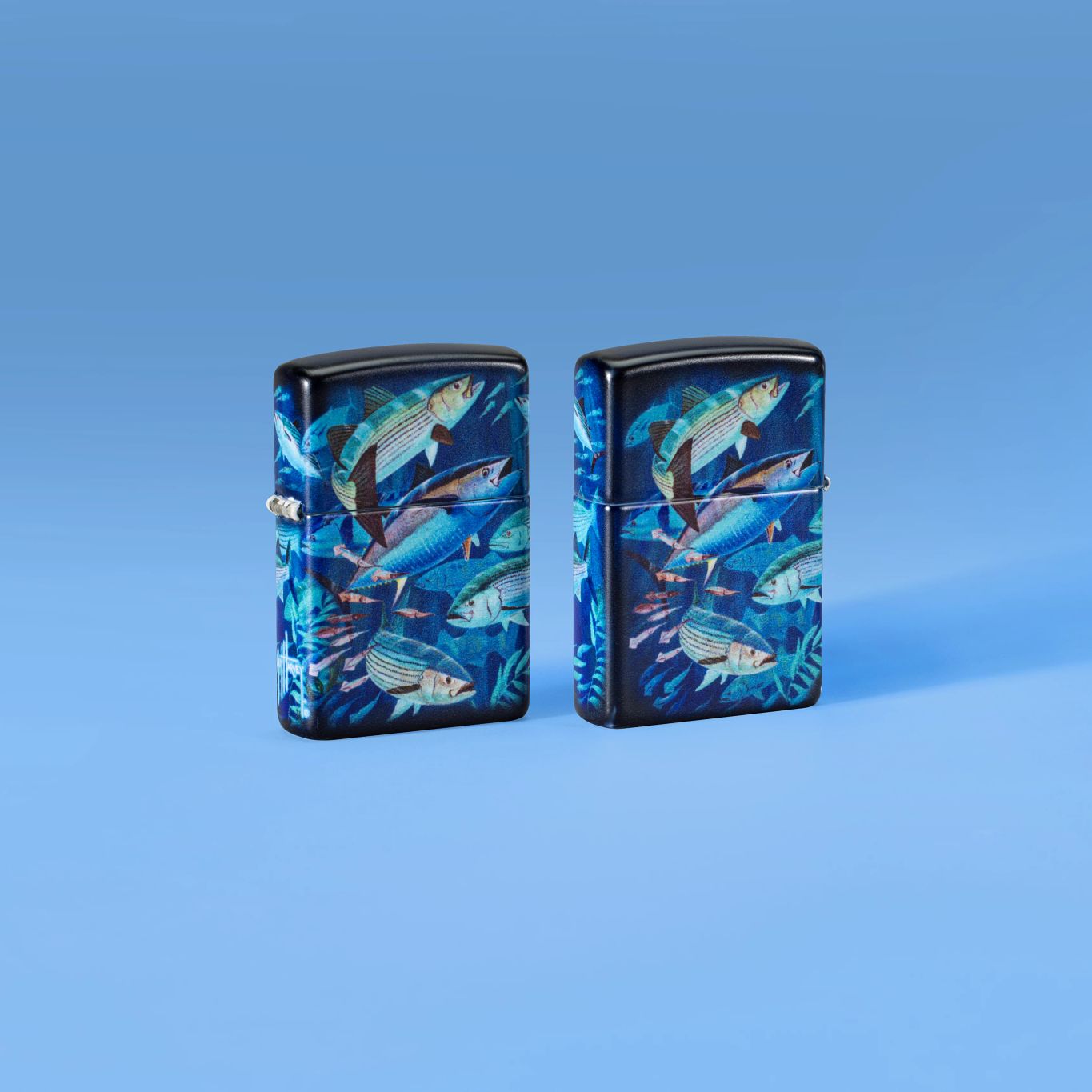 Lifestyle image of two Zippo Guy Harvey School of Fish Design White 540 Matte Windproof Lighters, one showing the front of the lighter and the other showing the back, standing on a pale blue background.