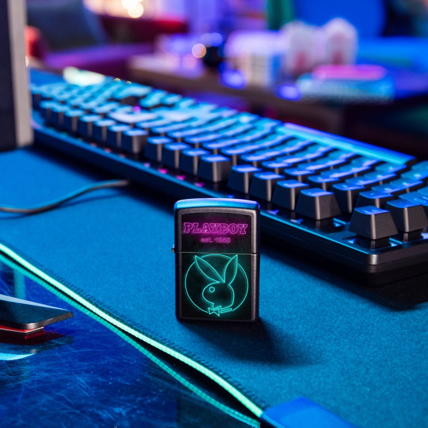 Lifestyle image of Zippo Playboy Neon Design Navy Matte Windproof Lighter standing in front of a keyboard with a blurred office setting with a blacklight in the background.