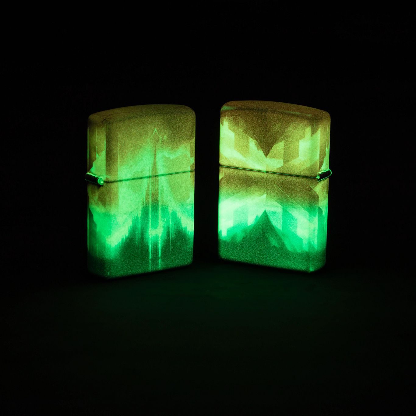 Lifestyle image of two Zippo Wicked City Design Glow in the dark Green Windproof Lighters; one showing the front of the lighter and the other showing the back and hinge side of the lighter, both glowing in the dark standing on a dark surface.
