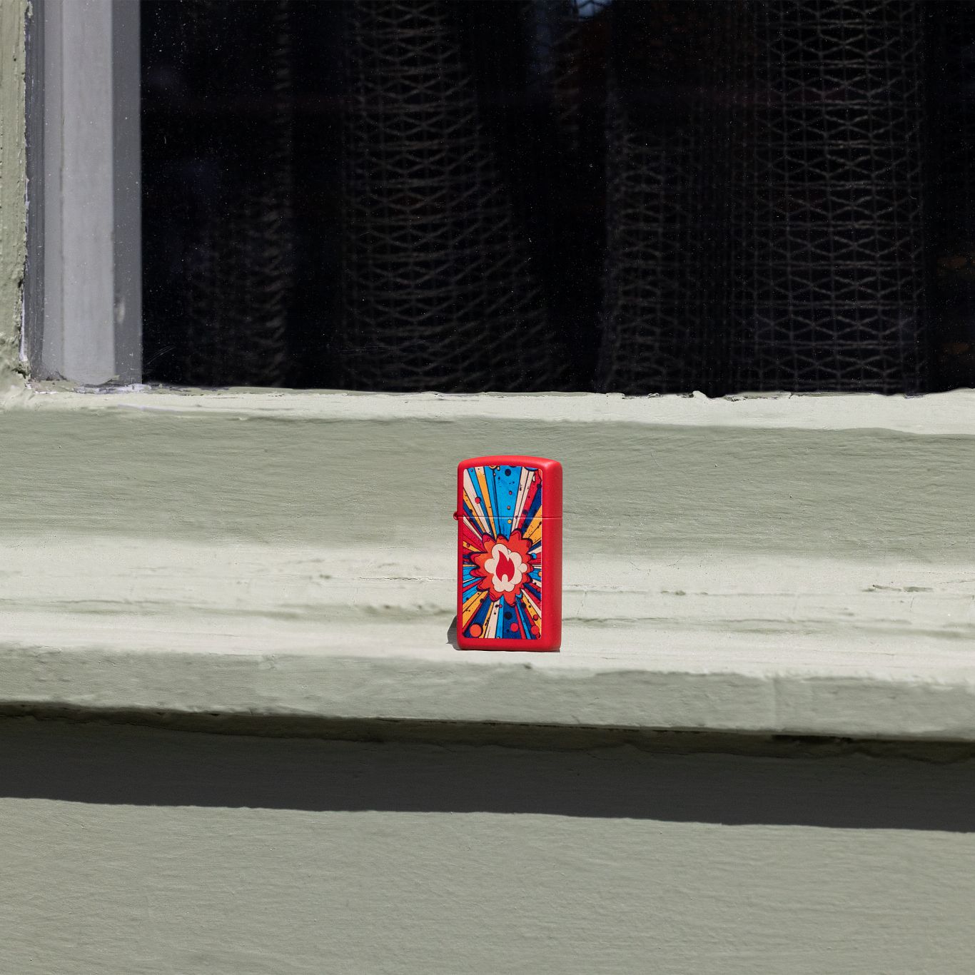 Lifestyle image of Zippo Comic Blast Design Slim Red Matte Windproof Lighter standing on a painted window ledge.
