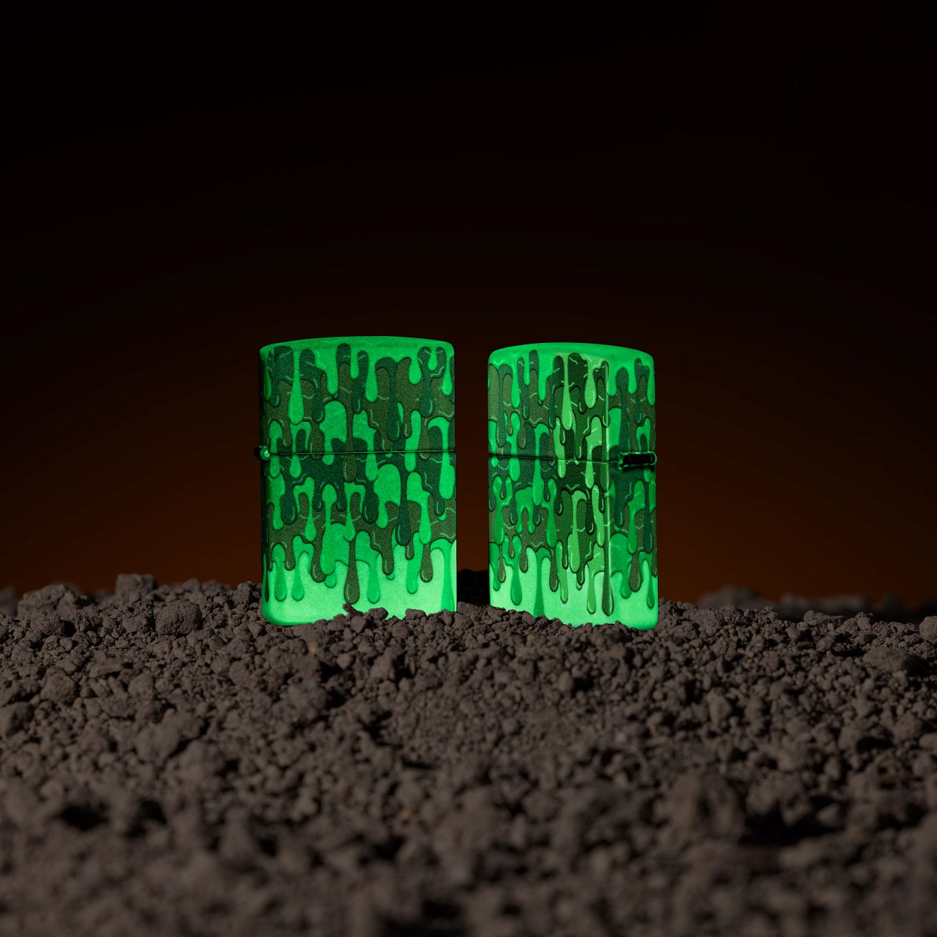 Lifestyle image of two Zippo Glowing Slime Design Glow in the Dark Windproof Lighters; one showing the front of the lighter and the other showing the back of the lighter, both glowing in the dark.