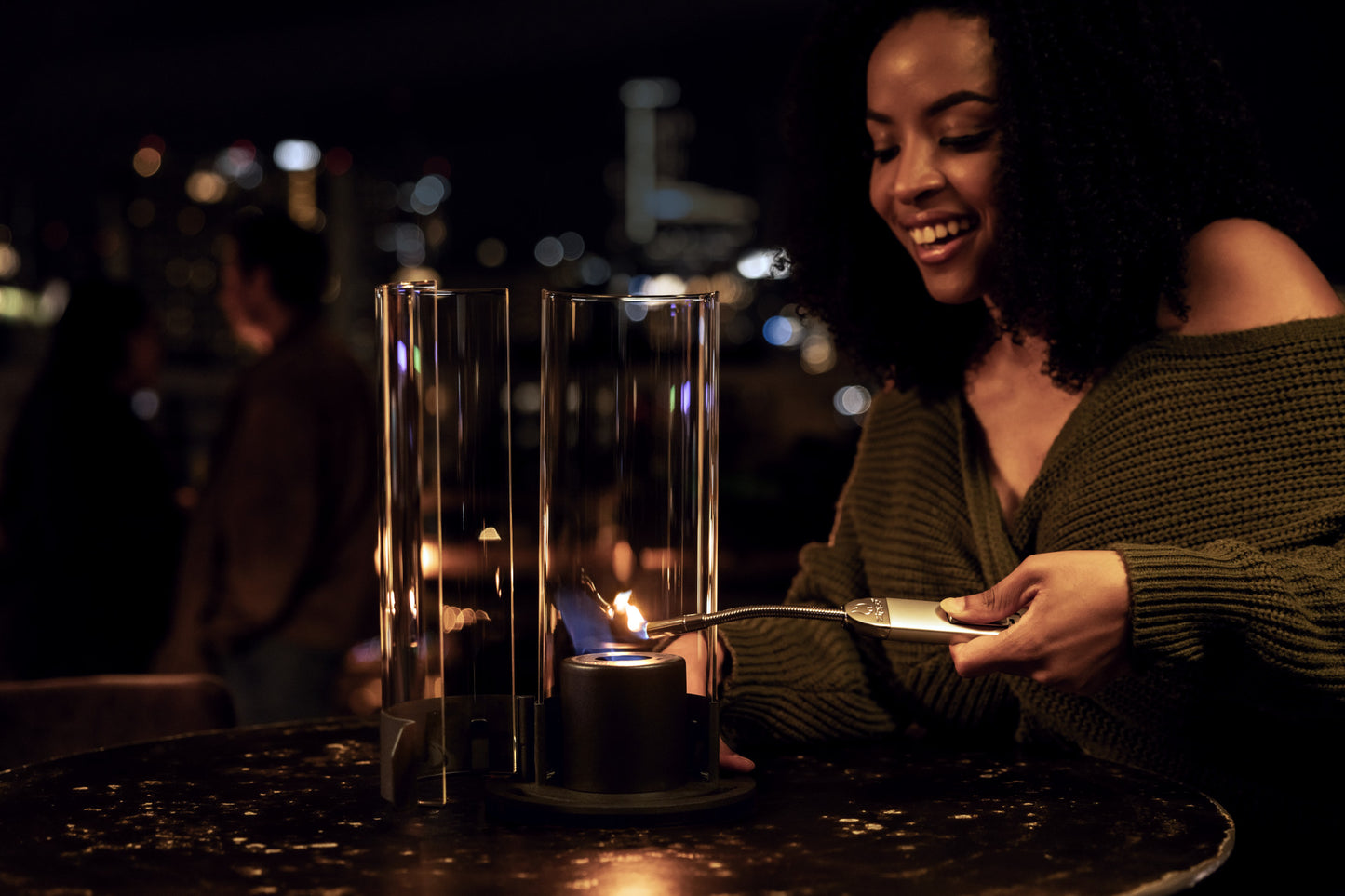 Lifestyle image of the Zippo FlameScapes® Spiral Fire Feature in black, lit on a table with a cityscape and couple in the background.