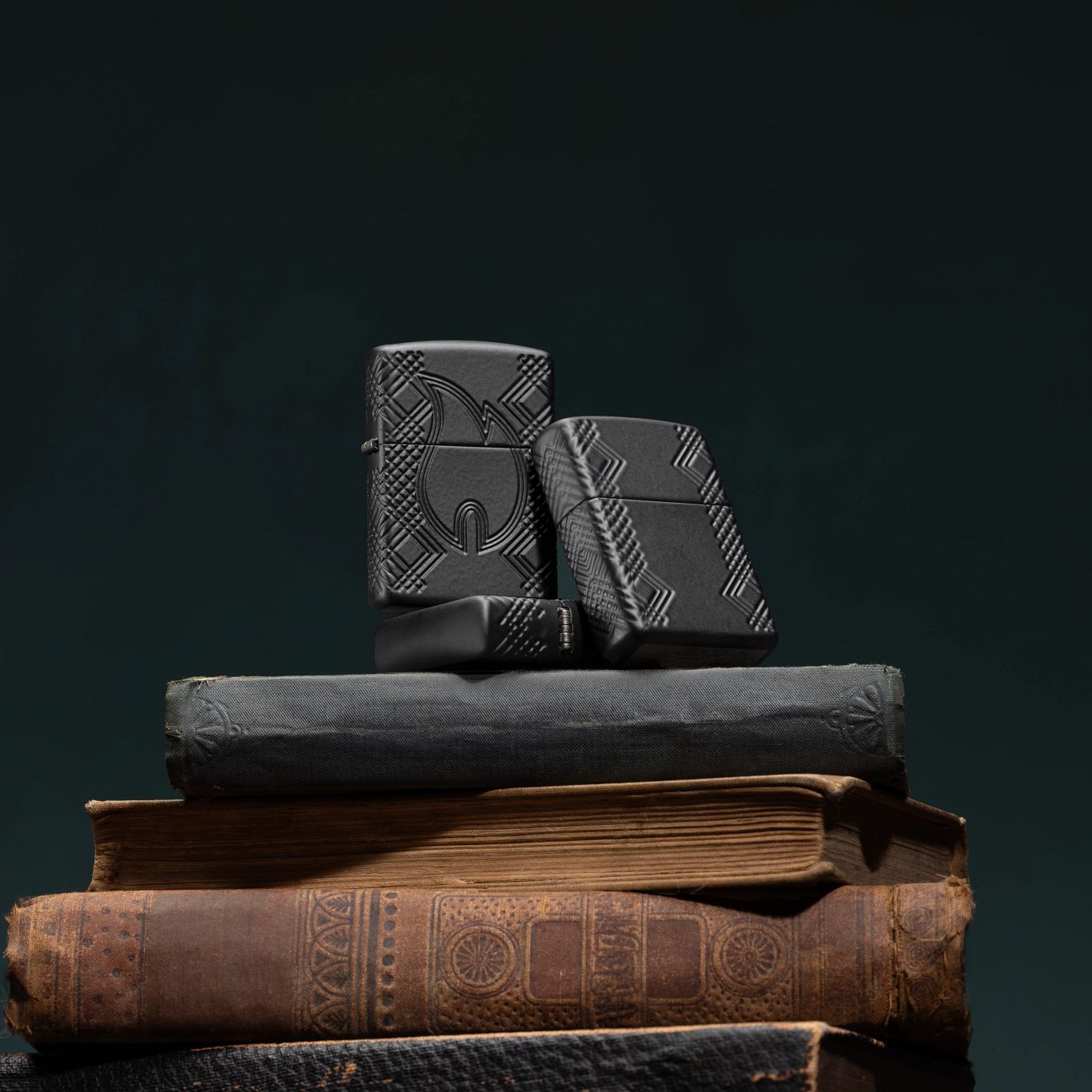 Lifestyle image of three Zippo Rivet Flame Design Armor® Black Matte Windproof Lighters on top of four stacked books with a dark grey background. One lighter is lying flat on top of a books showing the top of the lid and part of the hinge side of the lighter, the next standing on top of the lighter that is lying flat showing the front of the lighter, and the last lighter leaning against the first two lighters showing the back and the non-hinge side of the lighter.