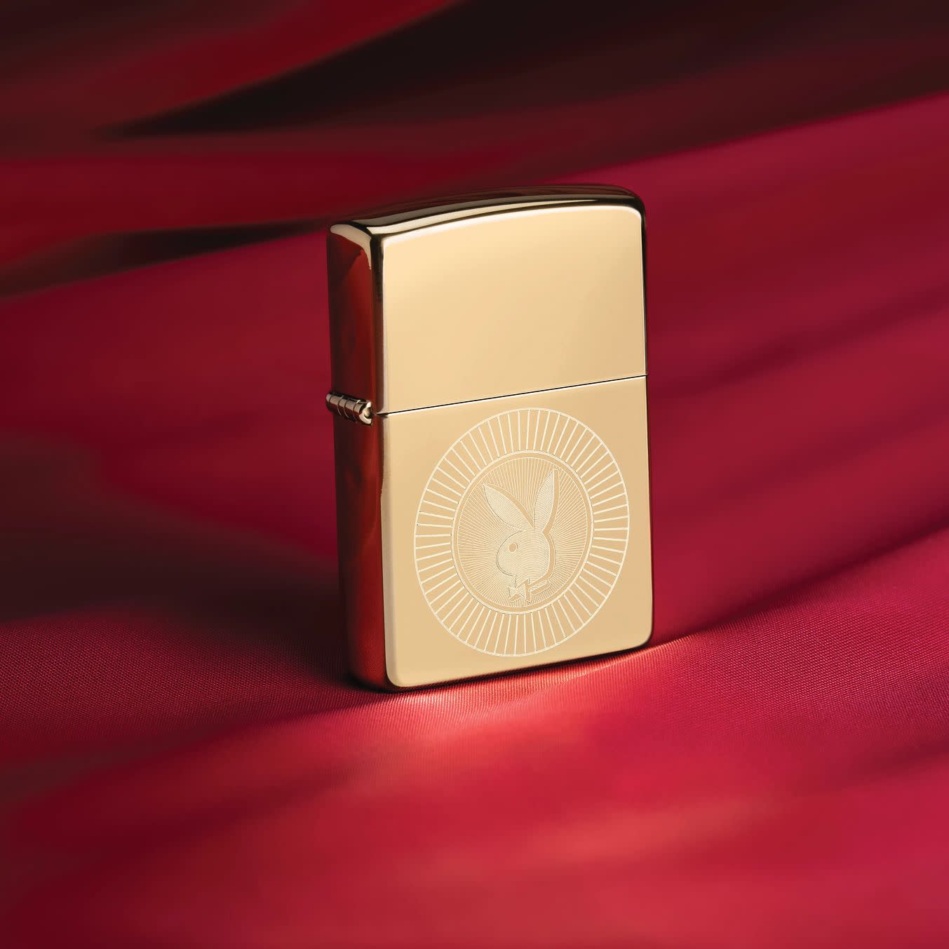 Lifestyle image of Zippo Playboy Rabbit Head Design High Polish Brass Windproof Lighter standing on a deep red fabric.