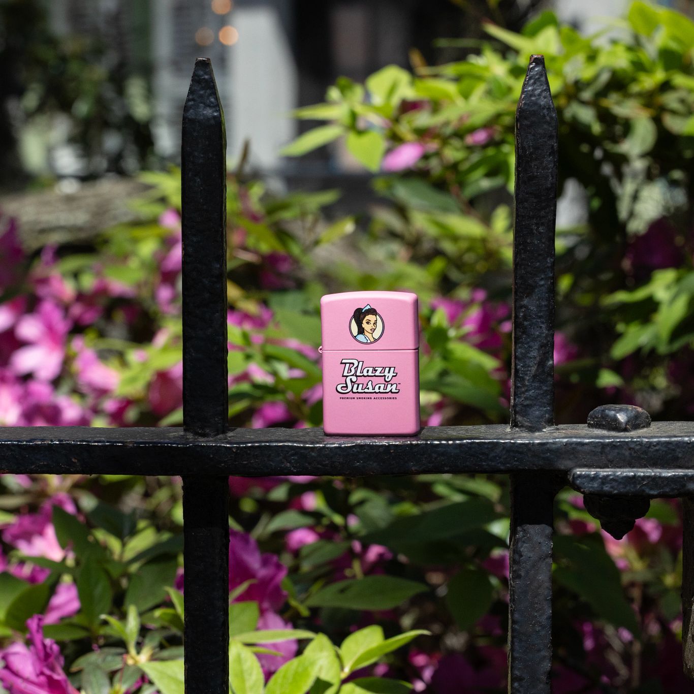 Lifestyle image of Zippo Blazy Susan™ Pink Logo Design Pink Matte Windproof Lighter standing on a wrought iron fence with blurred flowers in the background.