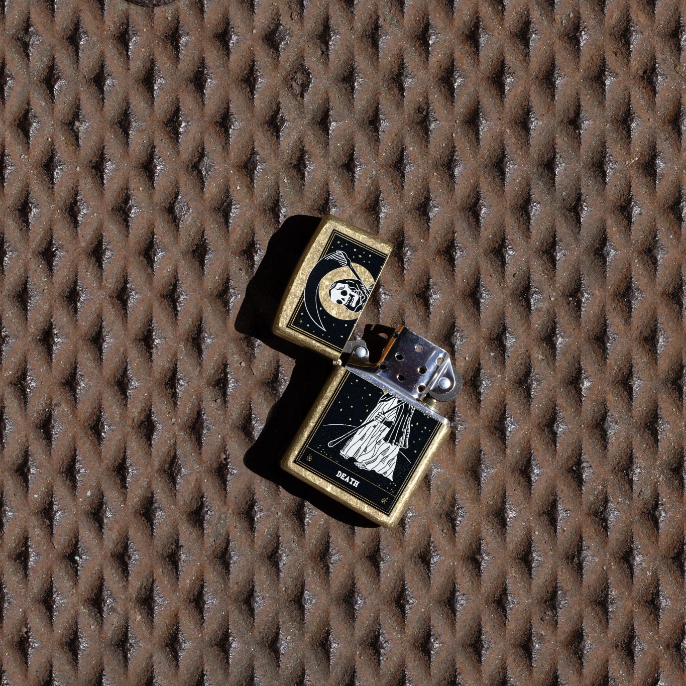Lifestyle image of Zippo Death Card Design Street Brass Windproof Lighter with its lid open and unlit laying on a rusted diamond tread surface.