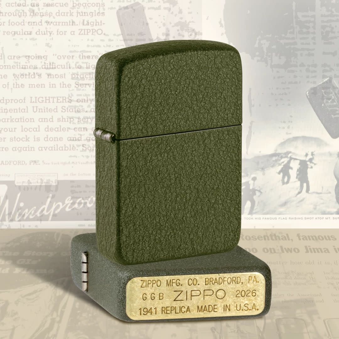 Lifestyle image of two Zippo 2026 Founder’s Day Commemorative Windproof Lighters; one lying flat showing the bottom stamp and hinge side of the lighter while the other lighter is standing on top of the lighter that is lying flat showing the front and hinge side of the lighter with a black and white poster as the background.