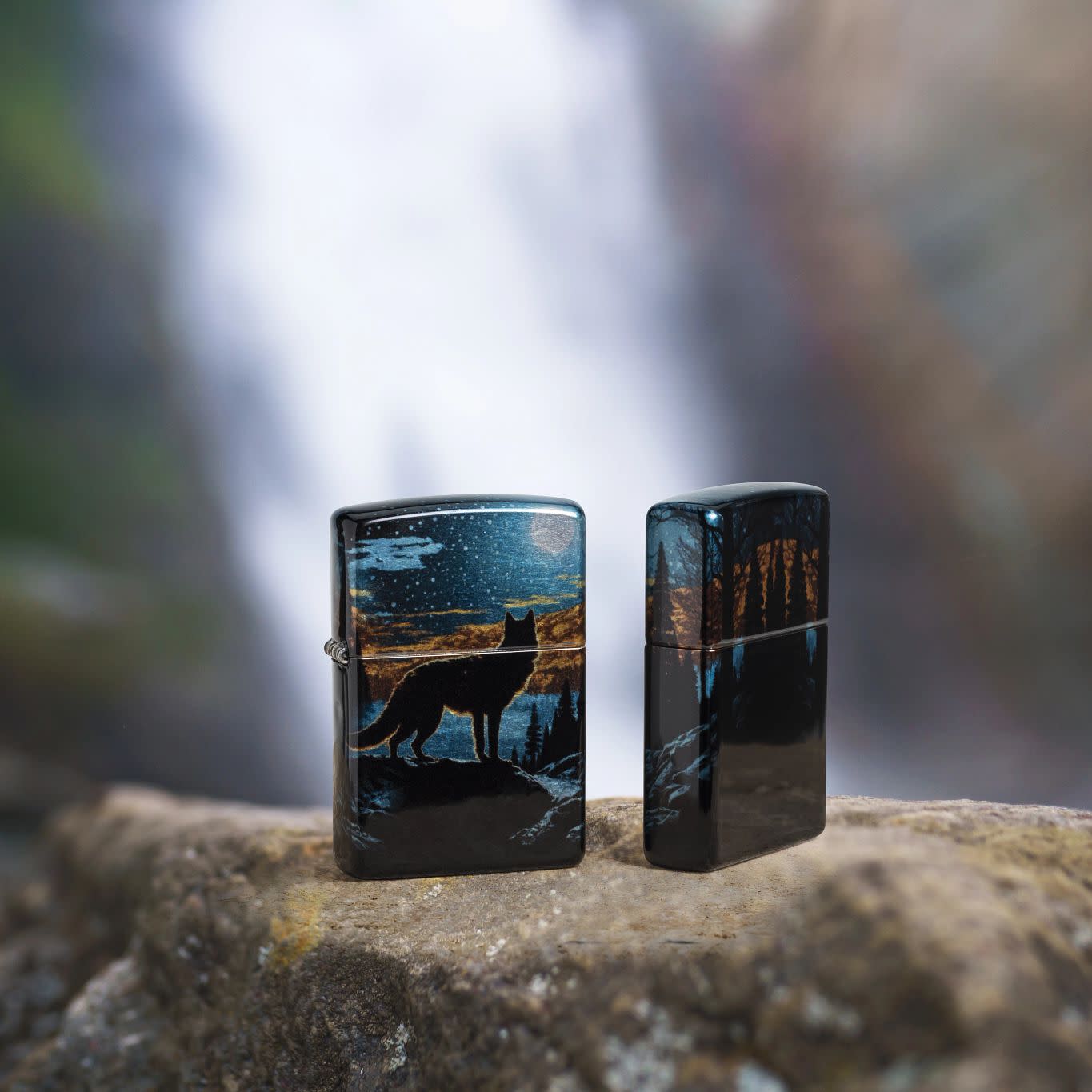 Lifestyle image of two Zippo Moonlight Wolf Design 540 Tumbled Chrome Windproof Lighters standing on a rock with a blurred waterfall in the background.