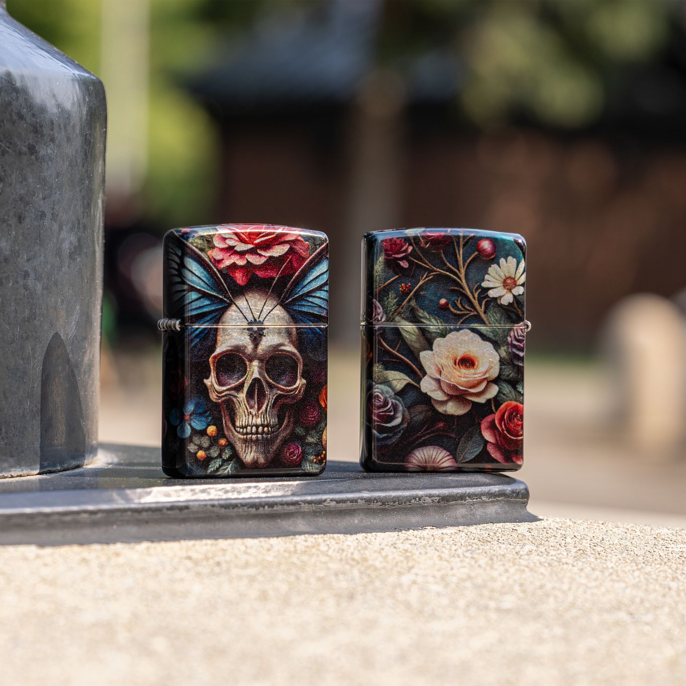 Lifestyle image of two Zippo Butterfly Skull Design 540 Tumbled Chrome Windproof Lighters; one showing the front and hinge side of the lighter and the other showing the back of the lighter, both standing on a metal structure with a concrete base. The background is an out of focus campus.