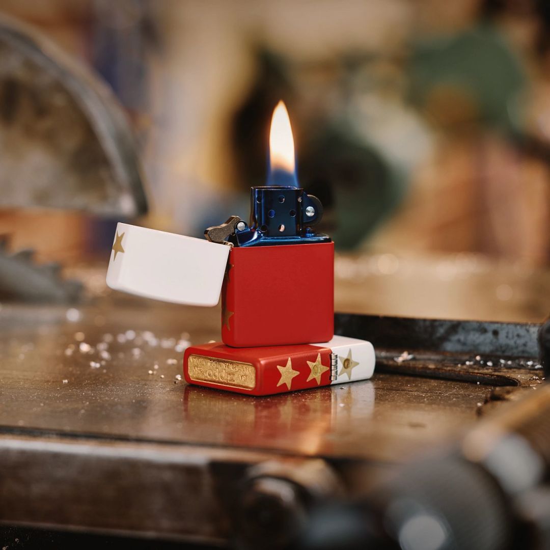Lifestyle image of two Zippo 250th Anniversary Special Edition White and Red Matte Windproof Lighters; one lying flat on a table saw workbench showing the hinge side and bottom stamp of the lighter while the other lighter is standing on top of the lighter that is lying flat, showing the front of the lighter open and lit.