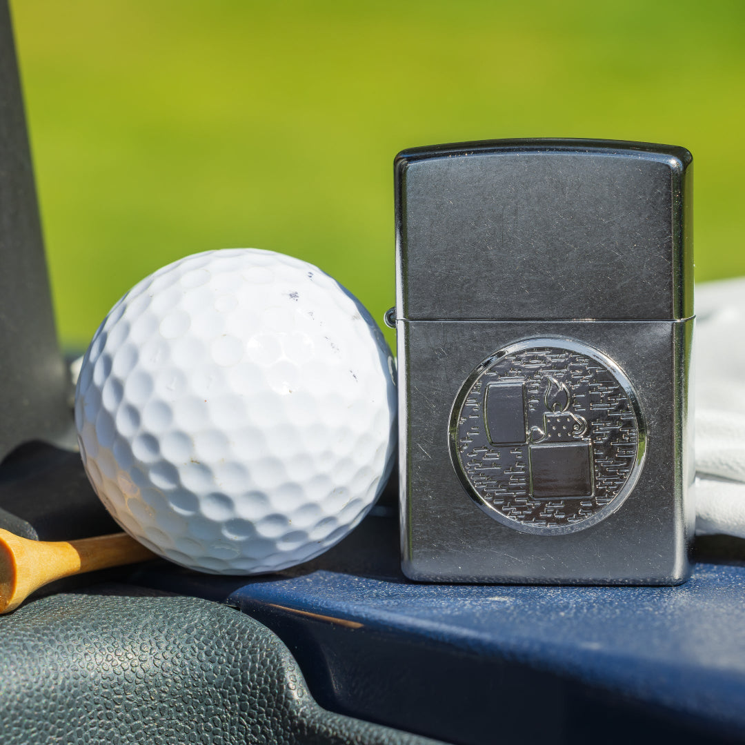 A Zippo lighter and golfball sitting on a blue surface with a green background.
