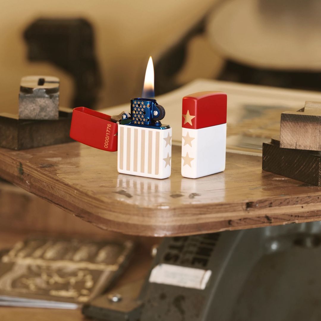 Lifestyle image of two Zippo 250th Anniversary Collectible Red and White Matte Windproof Lighters; one showing the front and non-hinge side of the lighter while open and lit and the other is showing the back and non-hinge side of the lighter while standing at a 3/4 angle. They are both standing on a wooden table next to some wooden blocks.