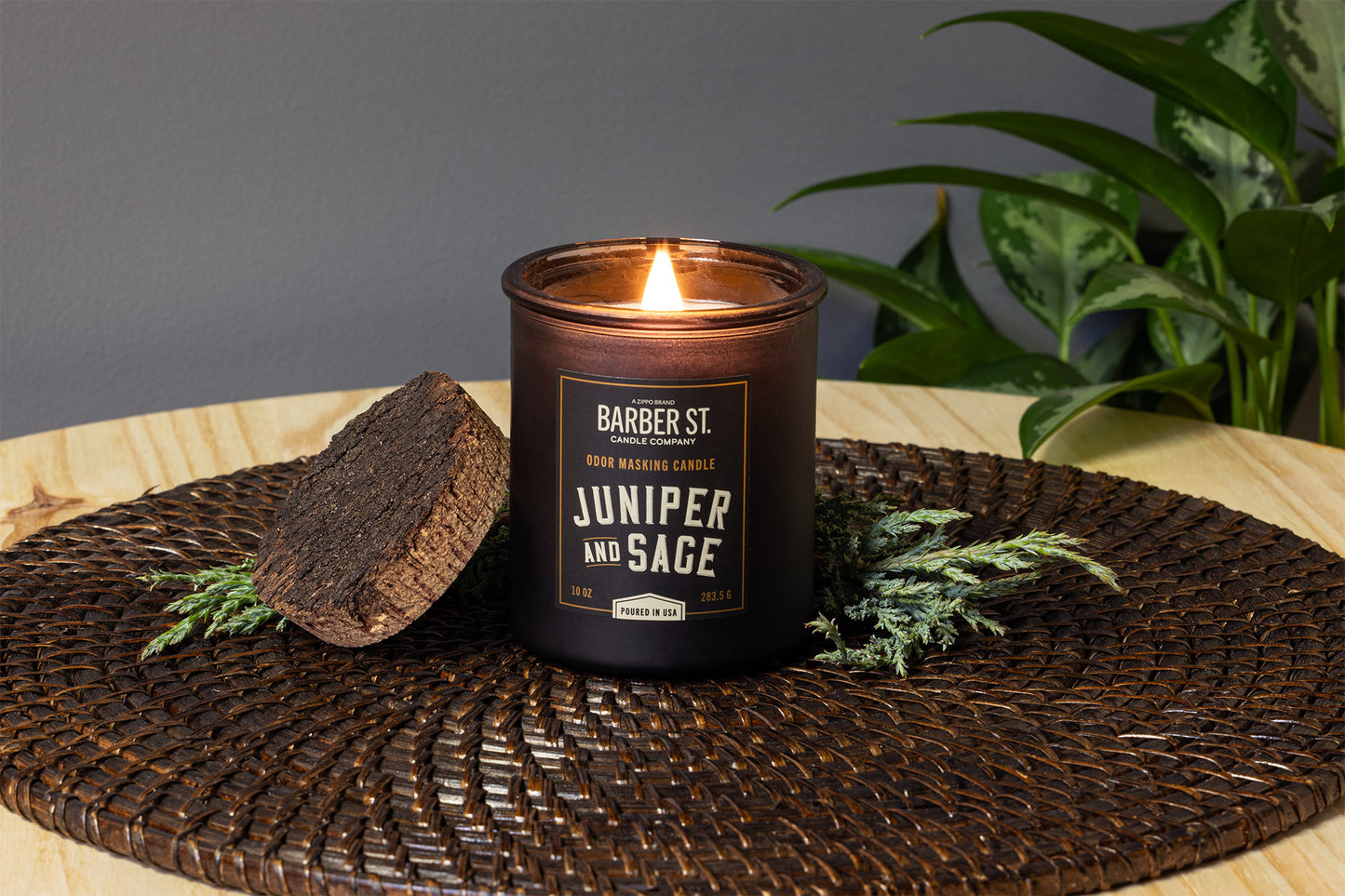 Lifestyle Image of Zippo Barber Street Candle Juniper and Sage on a wooden coffee table adorned with plants and brown sttiched table mat, lit.