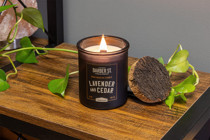 Lifestyle Image of Zippo Barber Street Candle Lavender and Cedar on a wooden end table with plant greenery, lit.