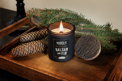 Lifestyle Image of Zippo Barber Street Candle Balsam and Fir on a wooden end table with pinecones and greenery, lit.