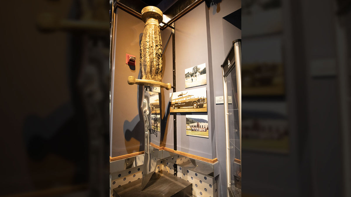 A large ornate Kodiak knife mounted vertically inside a glass display case, shown with framed pictures on the wall.