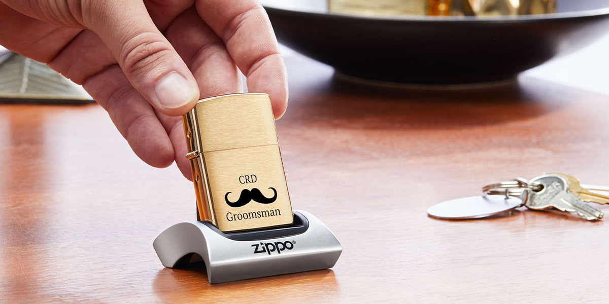 A hand holds a gold Zippo lighter engraved with 'CRD Groomsman' and a black mustache graphic, resting it on a silver Zippo display stand on a wooden table.