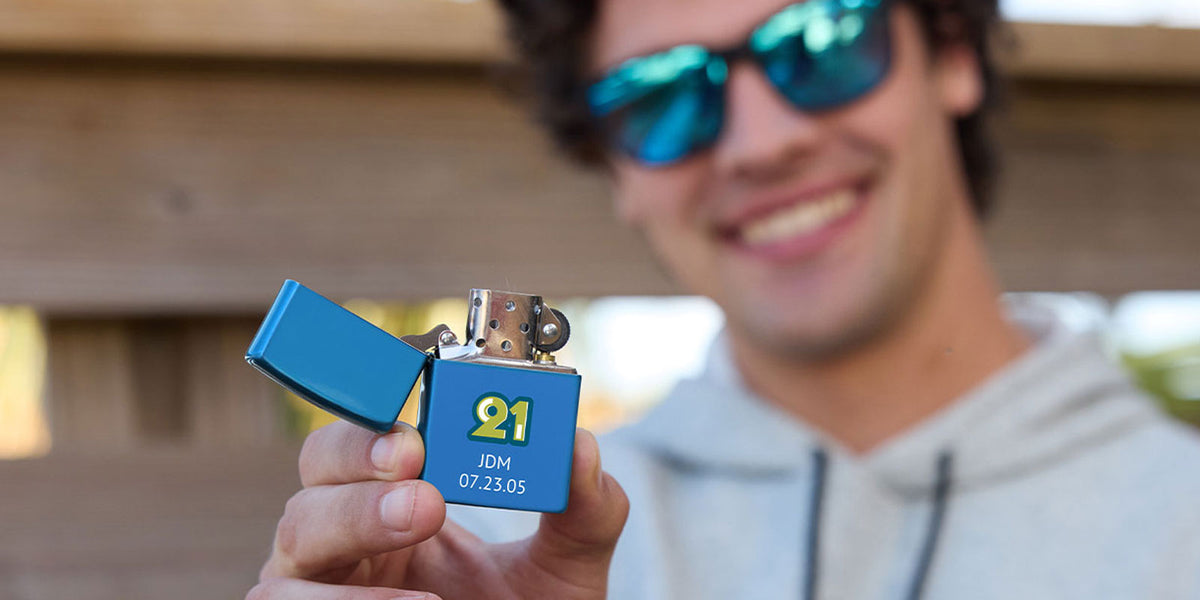 A person in a gray hoodie and blue mirrored sunglasses holds up a custom blue metal lighter with a yellow '21' design and date printed on the front, set against a blurred outdoor wooden background.